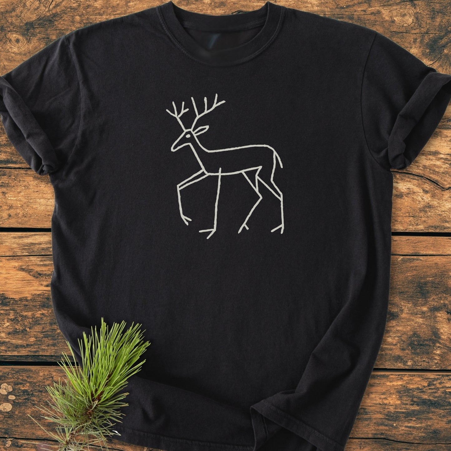 Deer Tribal - Vintage Wash Tee: A black T-shirt with a white line drawing of a stag with antlers, laid flat on wood, adorned with a small pine sprig for a rustic touch.