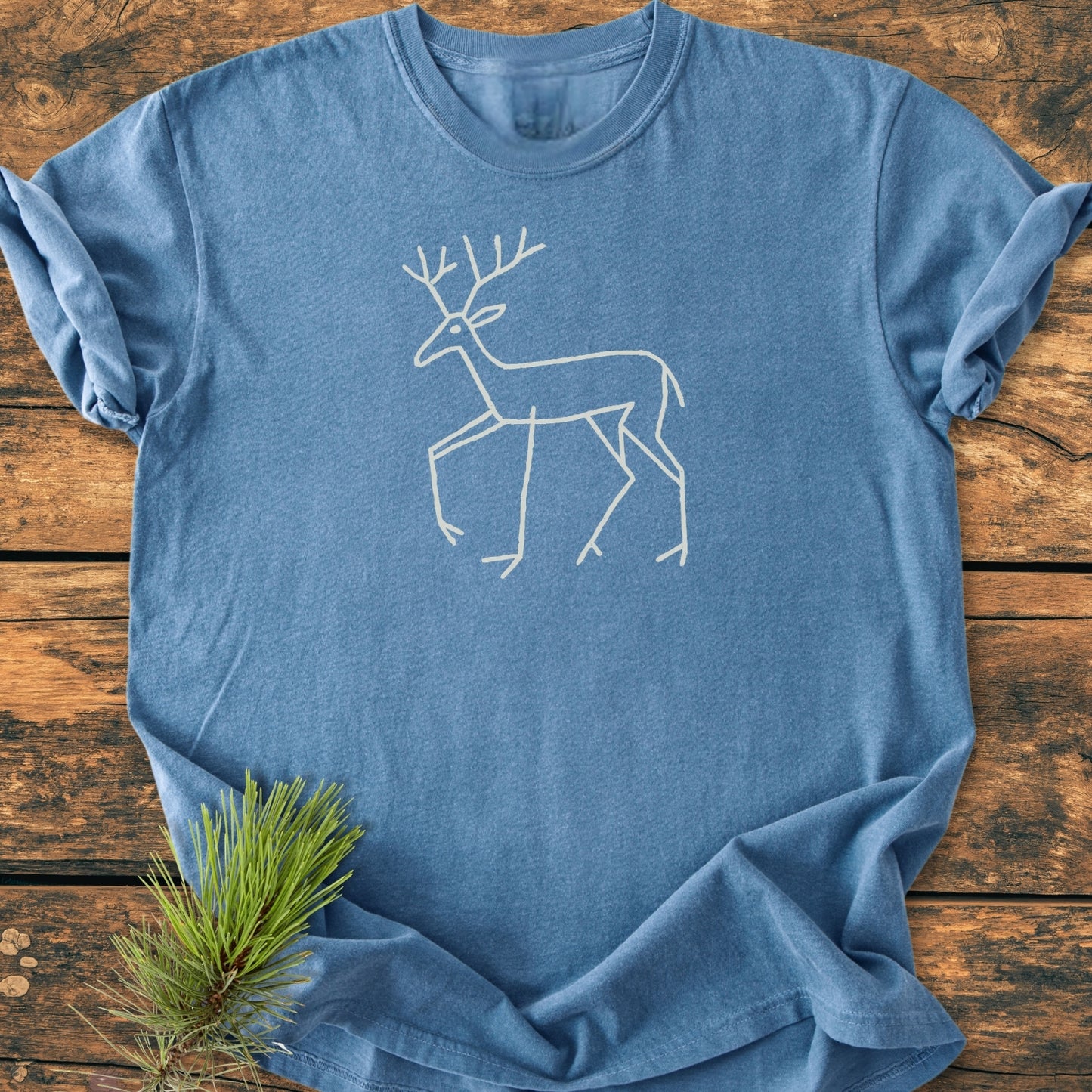 Deer Tribal - Vintage Wash Tee: A heather-blue short-sleeve T-shirt featuring a minimalist pale-cream line drawing of a deer with antlers, laid flat on a wooden surface with a pine sprig beside it.