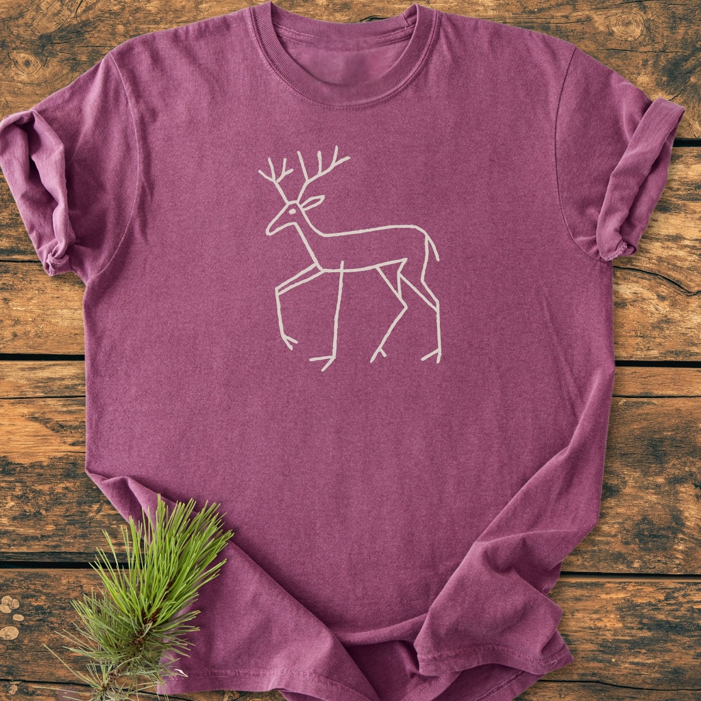 Deer Tribal - Vintage Wash Tee features a laid-flat design with a white deer line drawing on the front, set against a weathered wood surface with a pine sprig accent.