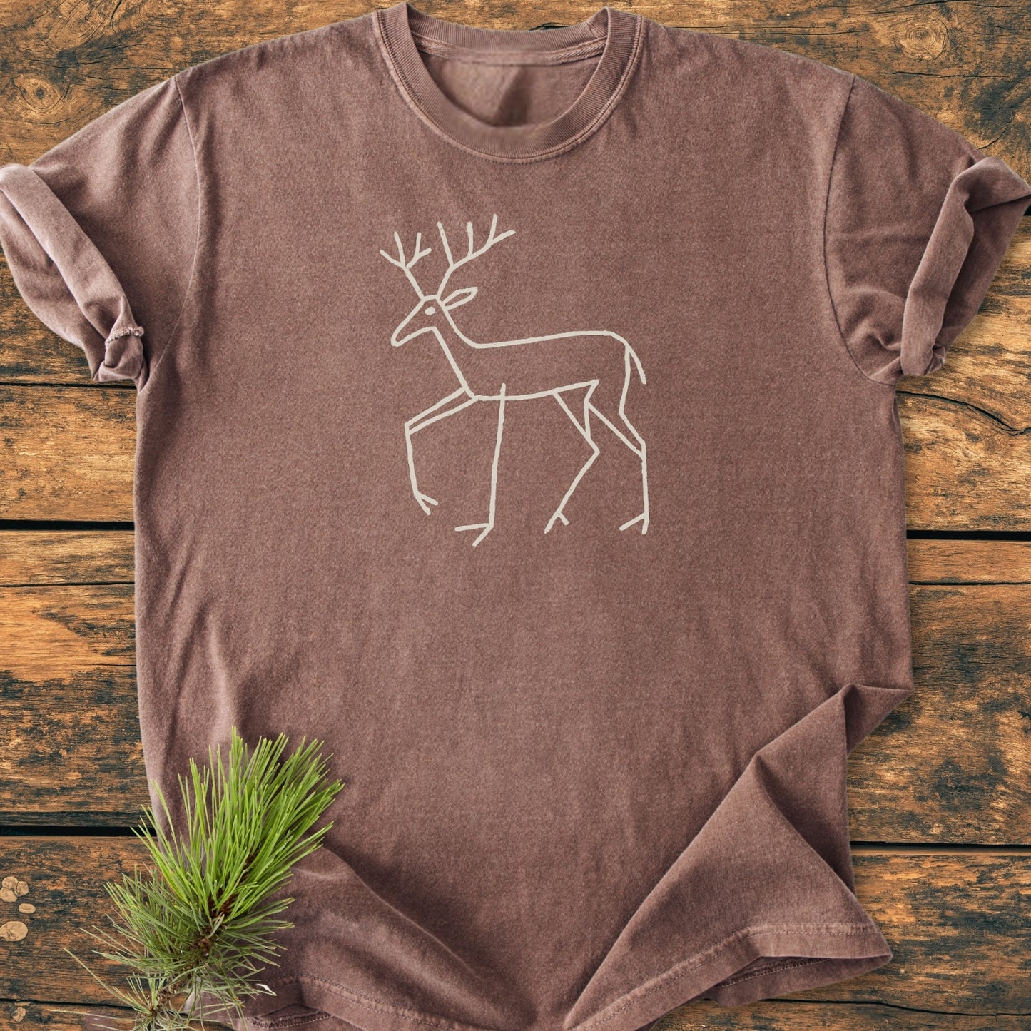 Deer Tribal - Vintage Wash Tee featuring a white line-drawing of a deer on a brown, stone-washed T-shirt, laid on rustic wood with a pine branch accent.