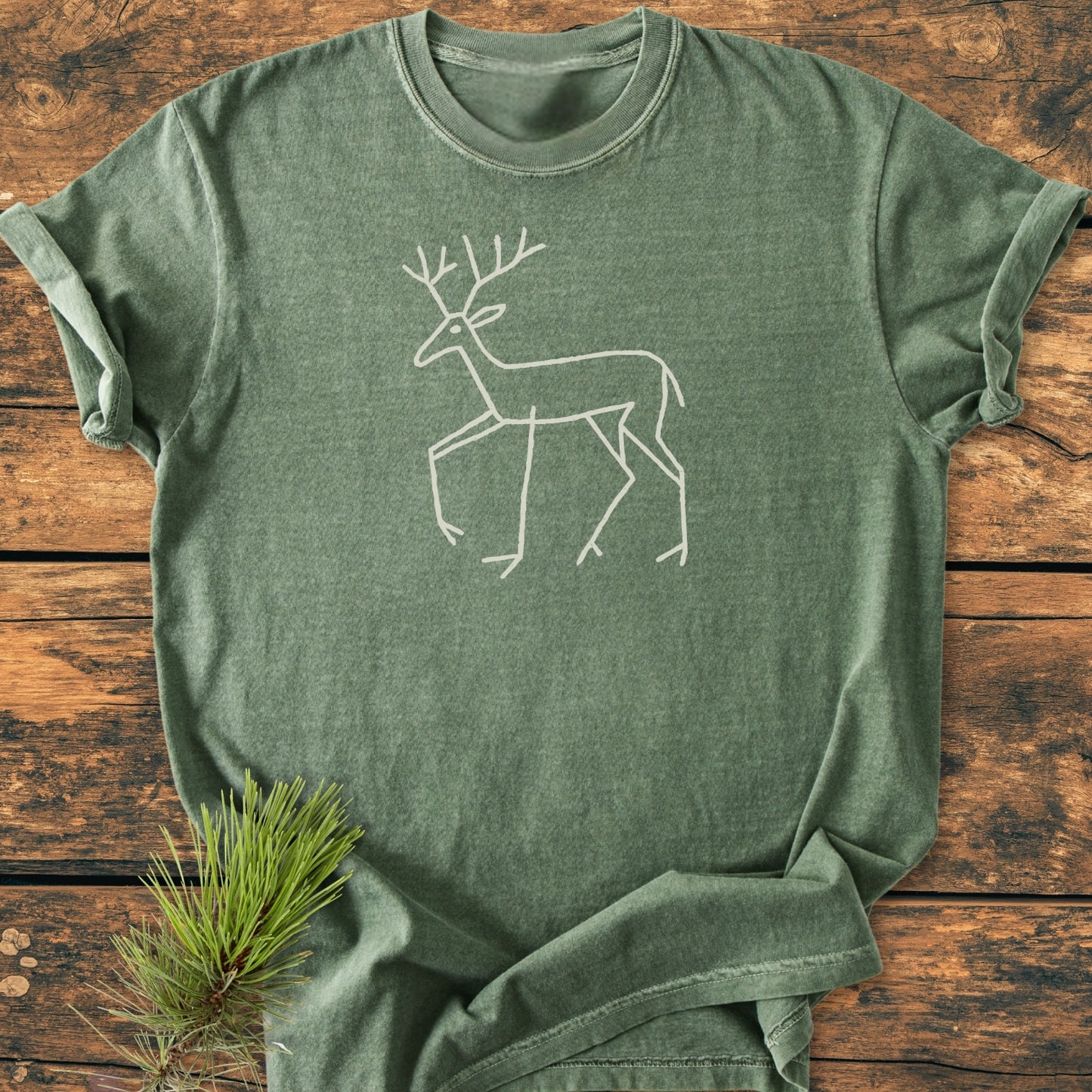 Deer Tribal - Vintage Wash Tee features an olive-green color with a white line drawing of a deer, laid on a wood surface. Includes a soft, relaxed fit, made from pre-shrunk ring-spun cotton.