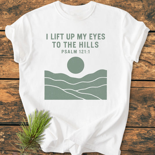 I Lift Up My Eyes - Vintage Wash Tee featuring a minimalist sage-green graphic of hills and sun on a white T-shirt, laid on a wooden surface with pine sprig accent.