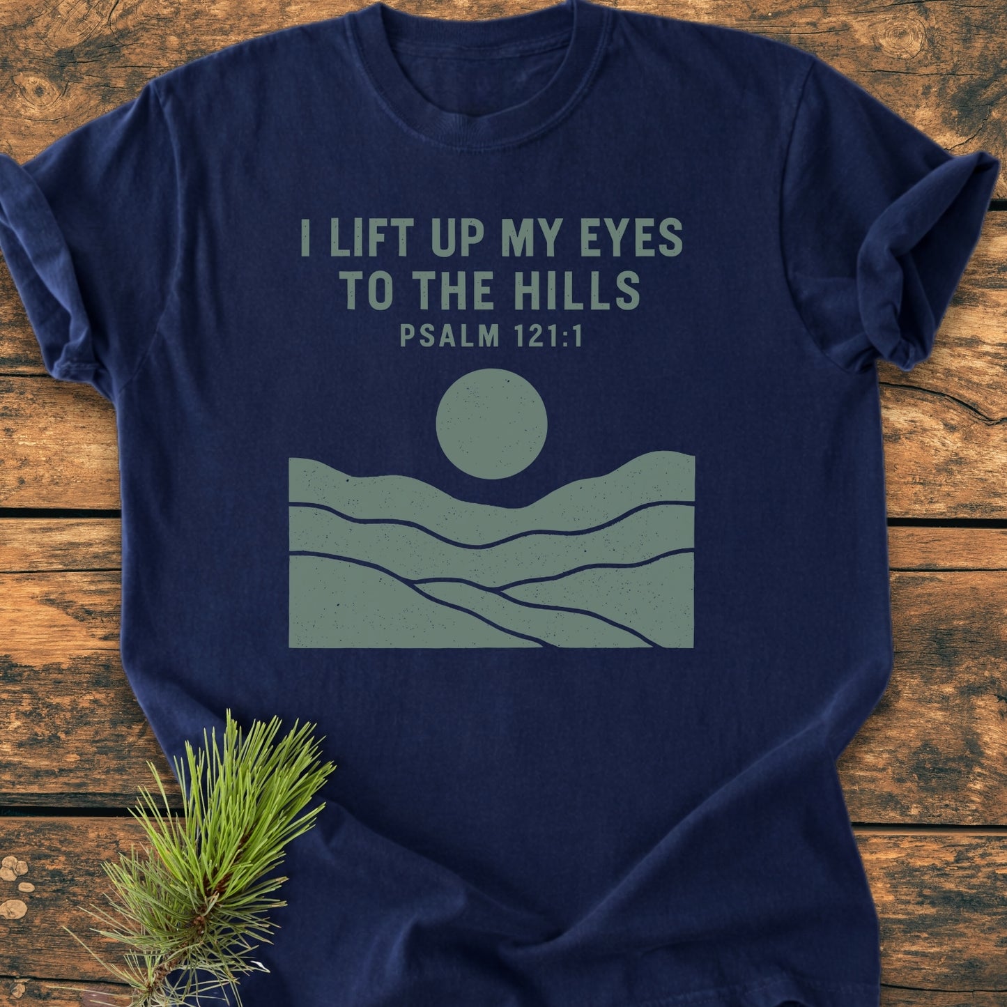 Navy-blue I Lift Up My Eyes - Vintage Wash Tee on wooden tabletop, featuring rolling hills, a sun graphic, and Psalm 121:1 text. Pine sprig beside the soft-washed, relaxed-fit cotton T-shirt.