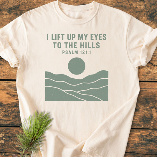 I Lift Up My Eyes - Vintage Wash Tee features Psalm 121:1 with rolling hills and a sun/moon graphic, displayed on a light-colored, soft-washed, garment-dyed T-shirt on a wooden surface.