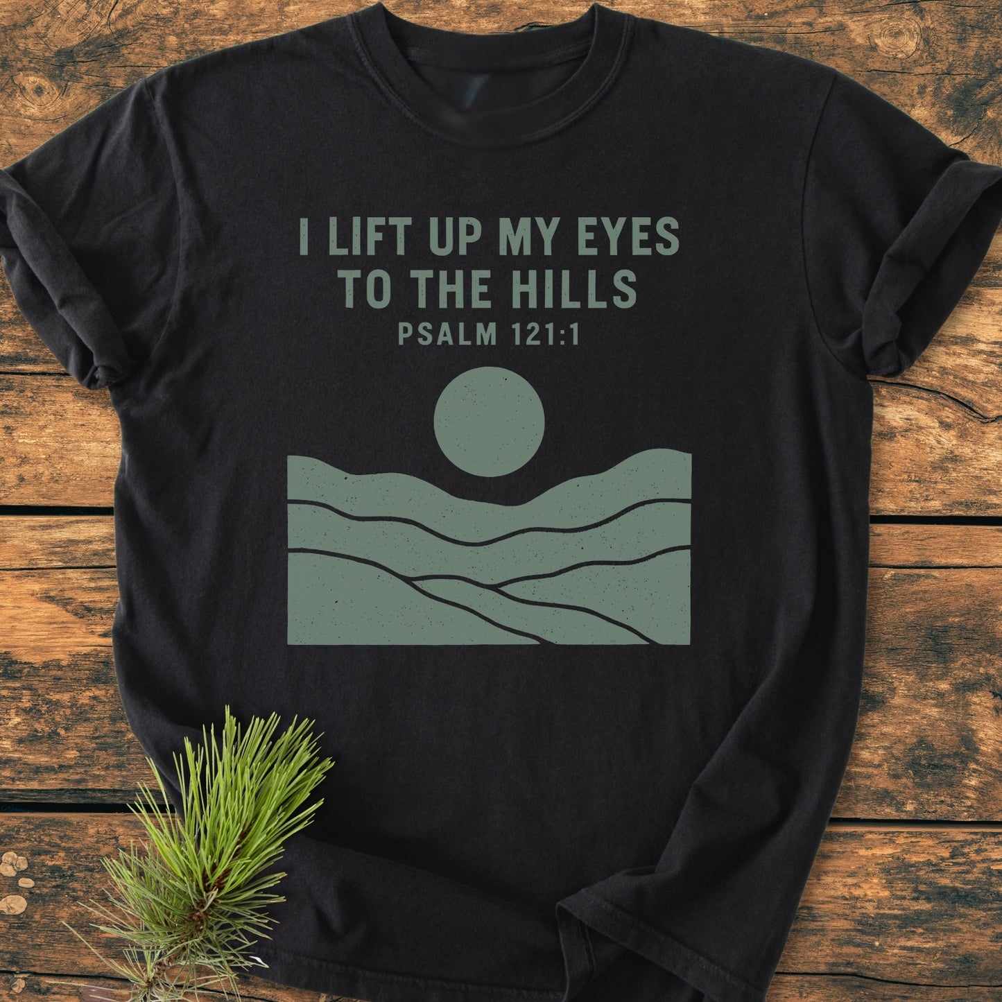 I Lift Up My Eyes - Vintage Wash Tee featuring Psalm 121:1 text in muted green with hills and sun graphic on black, displayed on rustic wood with a pine sprig beside.