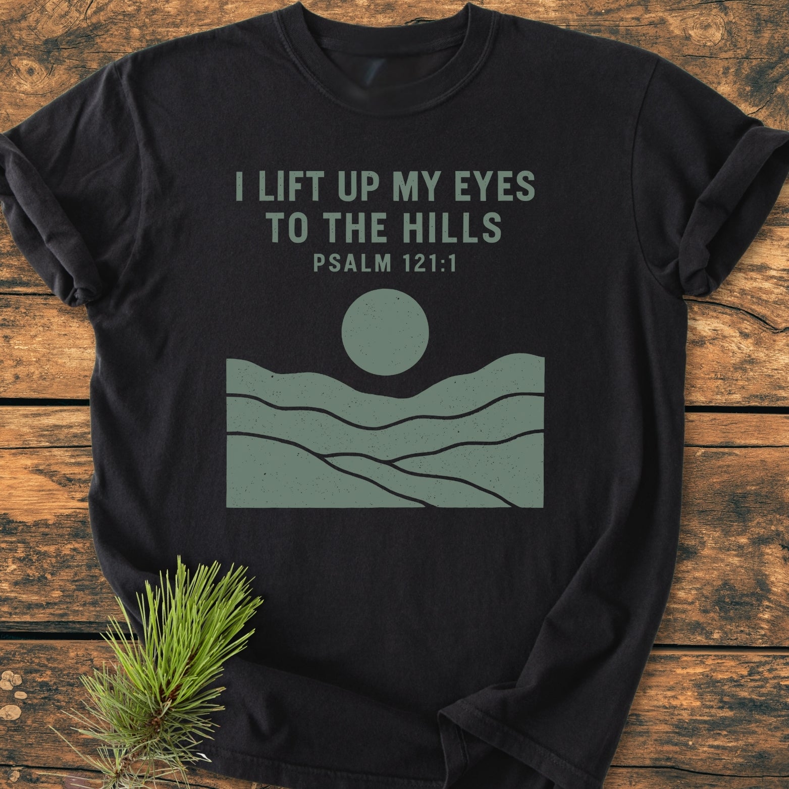I Lift Up My Eyes - Vintage Wash Tee featuring Psalm 121:1 text in muted green with hills and sun graphic on black, displayed on rustic wood with a pine sprig beside.