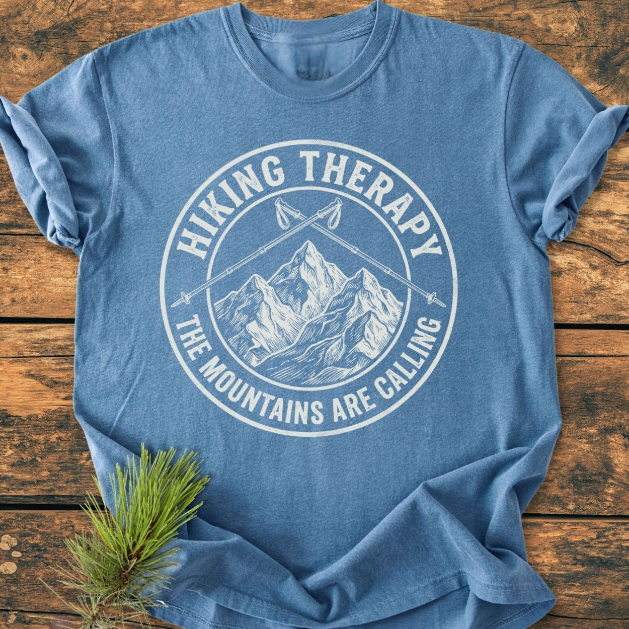 Hiking Therapy - Vintage Wash Tee featuring a circular mountain logo with crossed hiking poles, displayed on a wooden surface with a pine branch beside it, highlighting its relaxed fit and durable stitching.