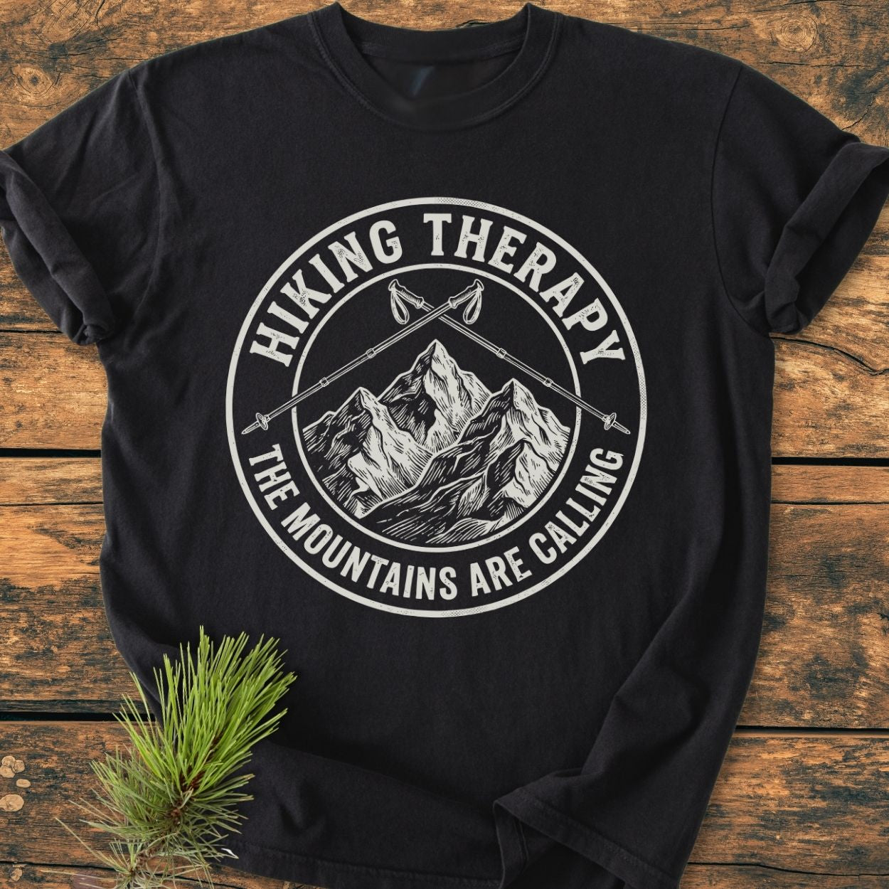 Hiking Therapy - Vintage Wash Tee features a vintage-style badge with mountain peaks and hiking poles, laid flat with a pine sprig on a wooden surface, showcasing its cozy, relaxed fit.