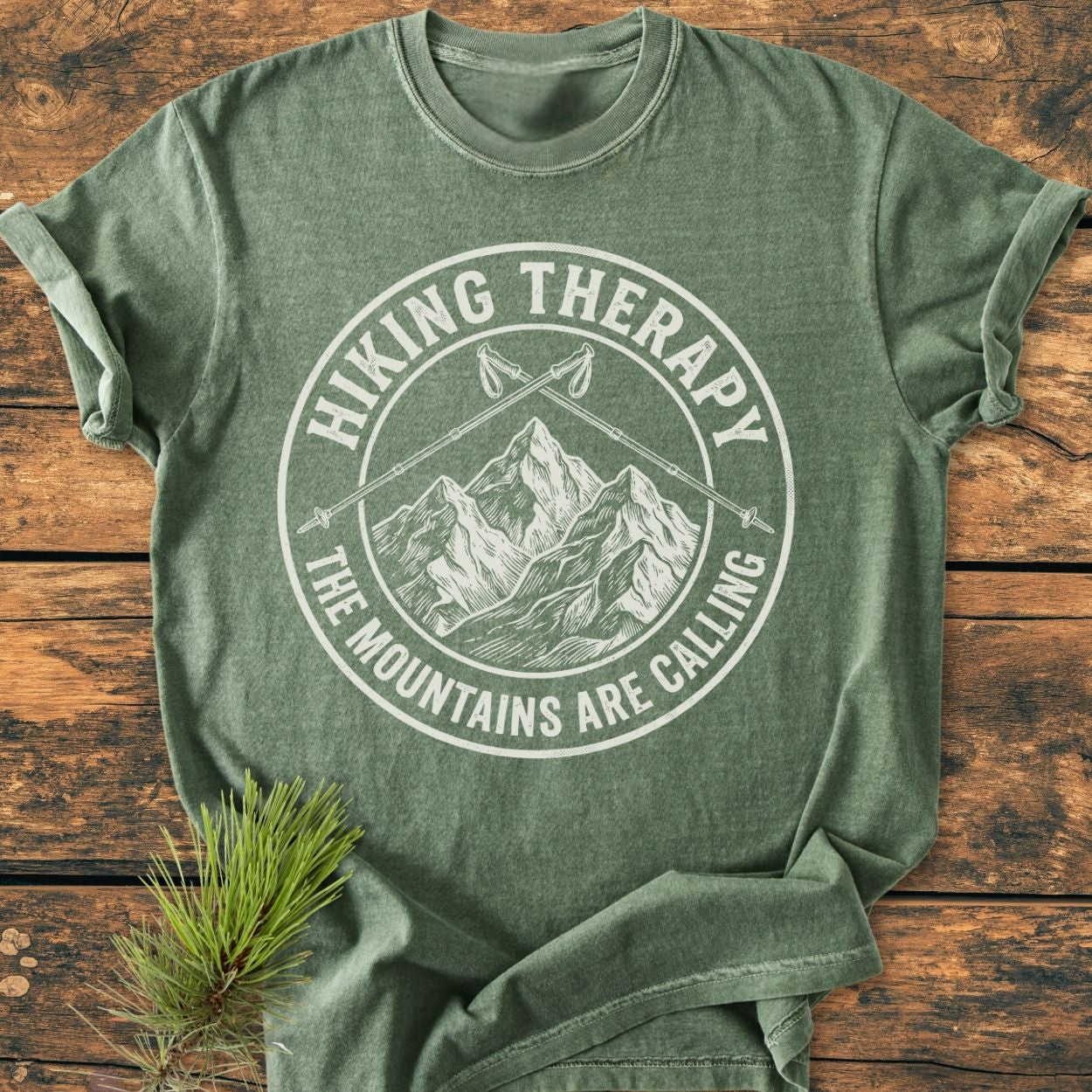 Hiking Therapy - Vintage Wash Tee, heather-green, features a white mountain graphic with hiking poles and motivational text, laid on wood. Includes pine sprig accent, emphasizing outdoor adventure spirit.