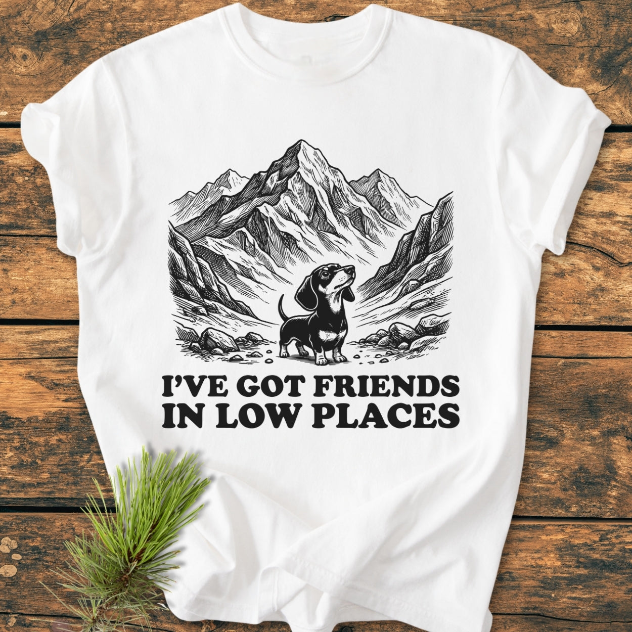 Friends In Low Places - Vintage Wash Tee features a stylized mountain range with a dachshund and punny slogan, laid on wood, adorned with a pine sprig. 