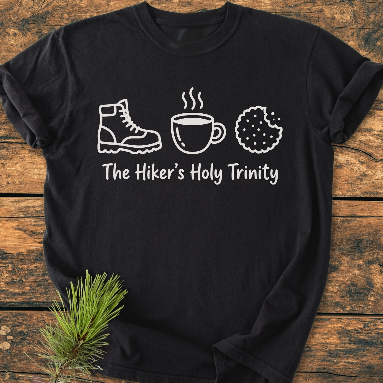 The Hiker's Holy Trinity - Vintage Wash Tee features line drawings of a boot, mug, and cookie on black fabric. The soft, relaxed-fit tee is laid on a wooden surface with a pine sprig beside it.