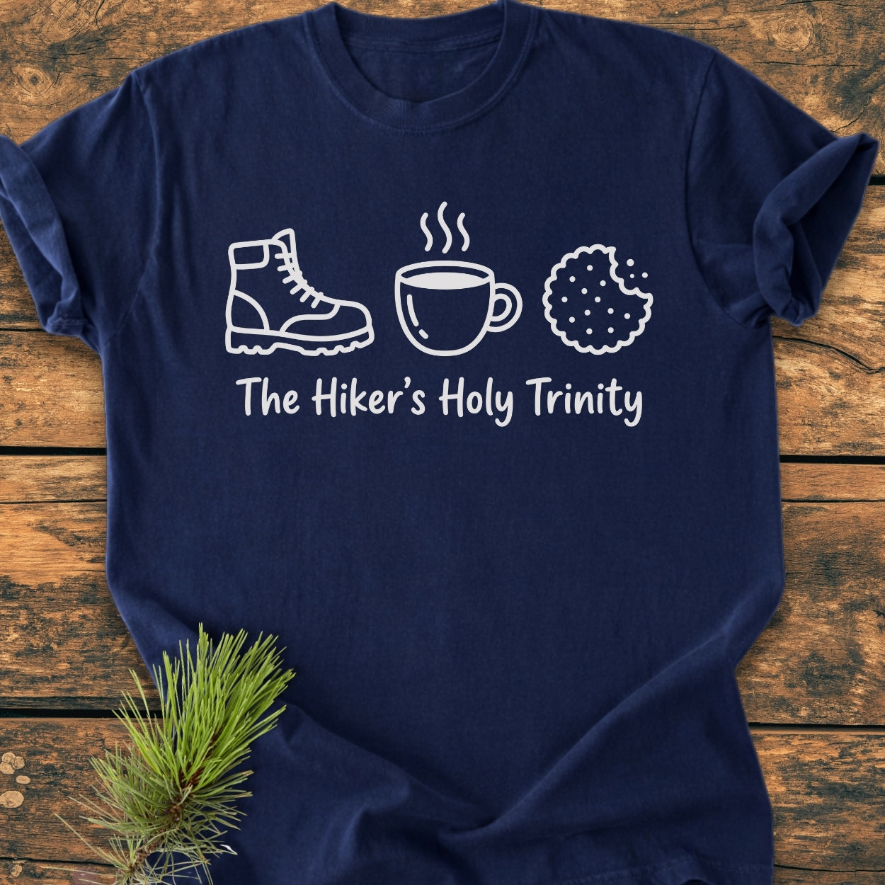 The Hiker's Holy Trinity - Vintage Wash Tee features white line-drawings of a boot, mug, and cookie with the caption on a soft, navy garment-dyed T-shirt laid on a wood surface.