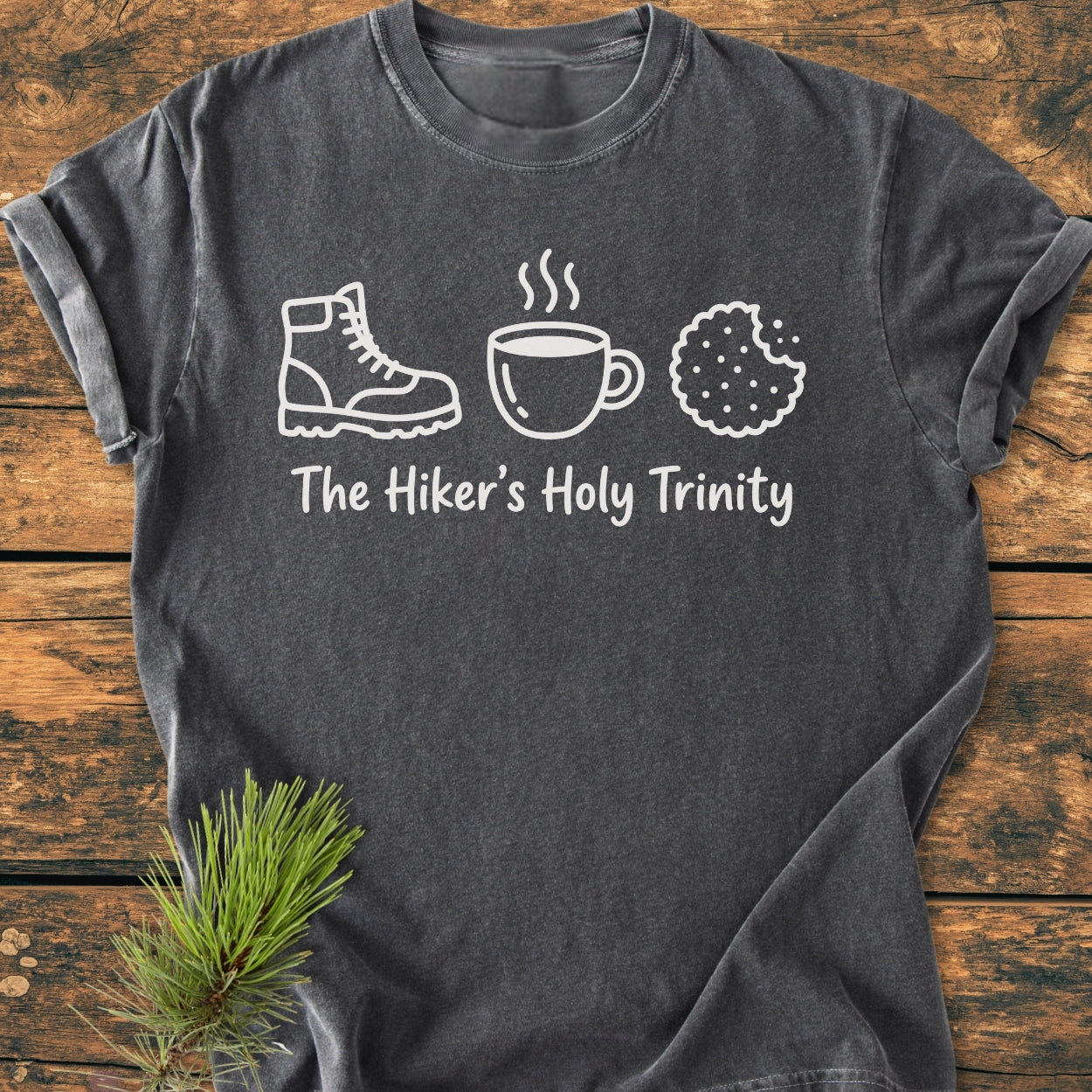 The Hiker's Holy Trinity - Vintage Wash Tee features a dark-gray design with white line drawings of a boot, mug, and cookie. Text reads “The Hiker’s Holy Trinity” on soft, garment-dyed cotton.