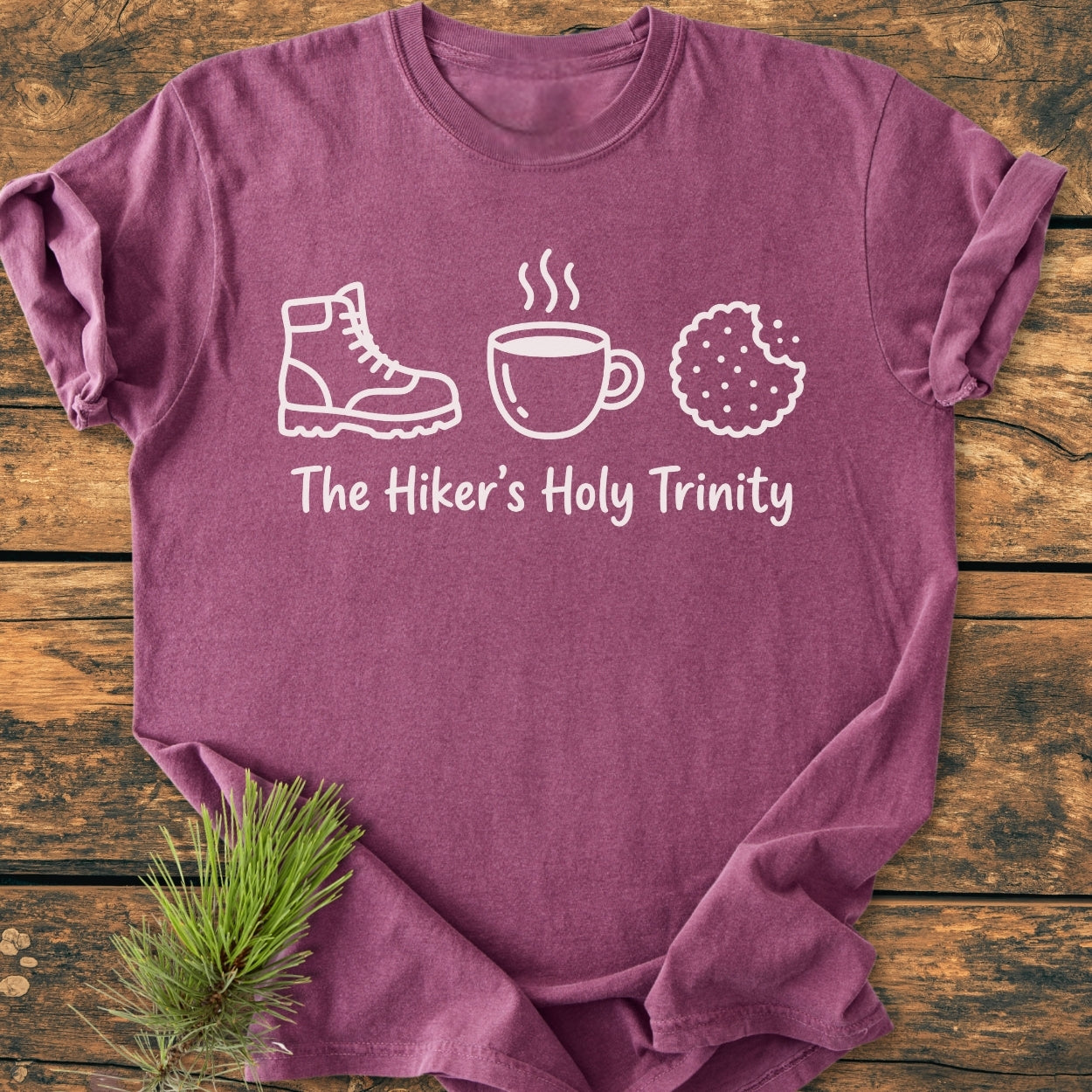 The Hiker's Holy Trinity - Vintage Wash Tee features icons of a hiking boot, mug, and cookie with the phrase below, laid on rustic wood with a pine sprig accent.