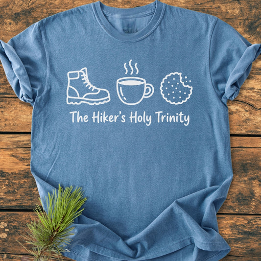 Heather-blue The Hiker's Holy Trinity tee featuring icons of a boot, mug, and cookie, with a pine sprig accent, laid on wood. Cozy, durable, 100% cotton, relaxed fit.