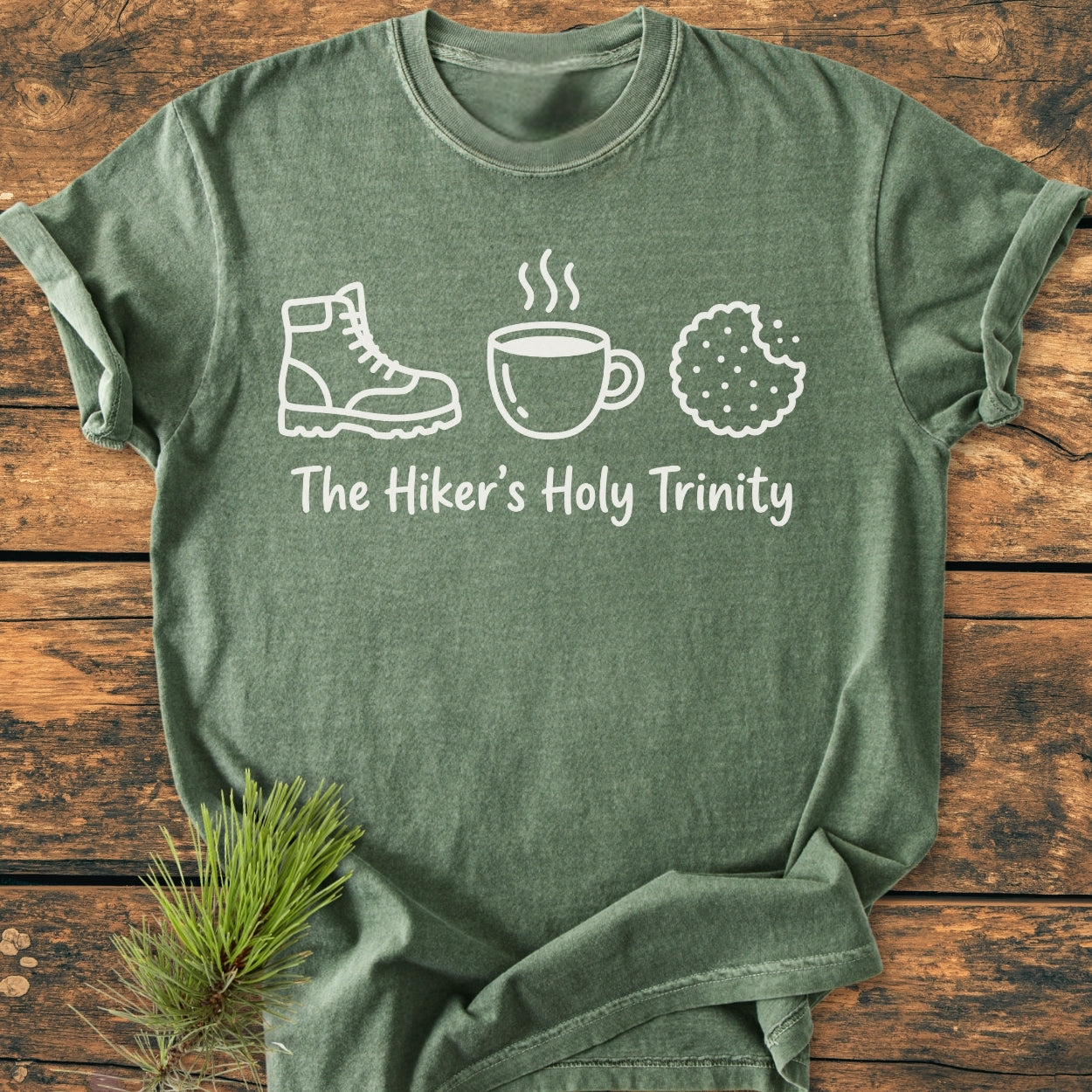 The Hiker's Holy Trinity - Vintage Wash Tee features icons of a boot, mug, and cookie in white on an olive-green shirt, laid flat with a pine sprig accent.