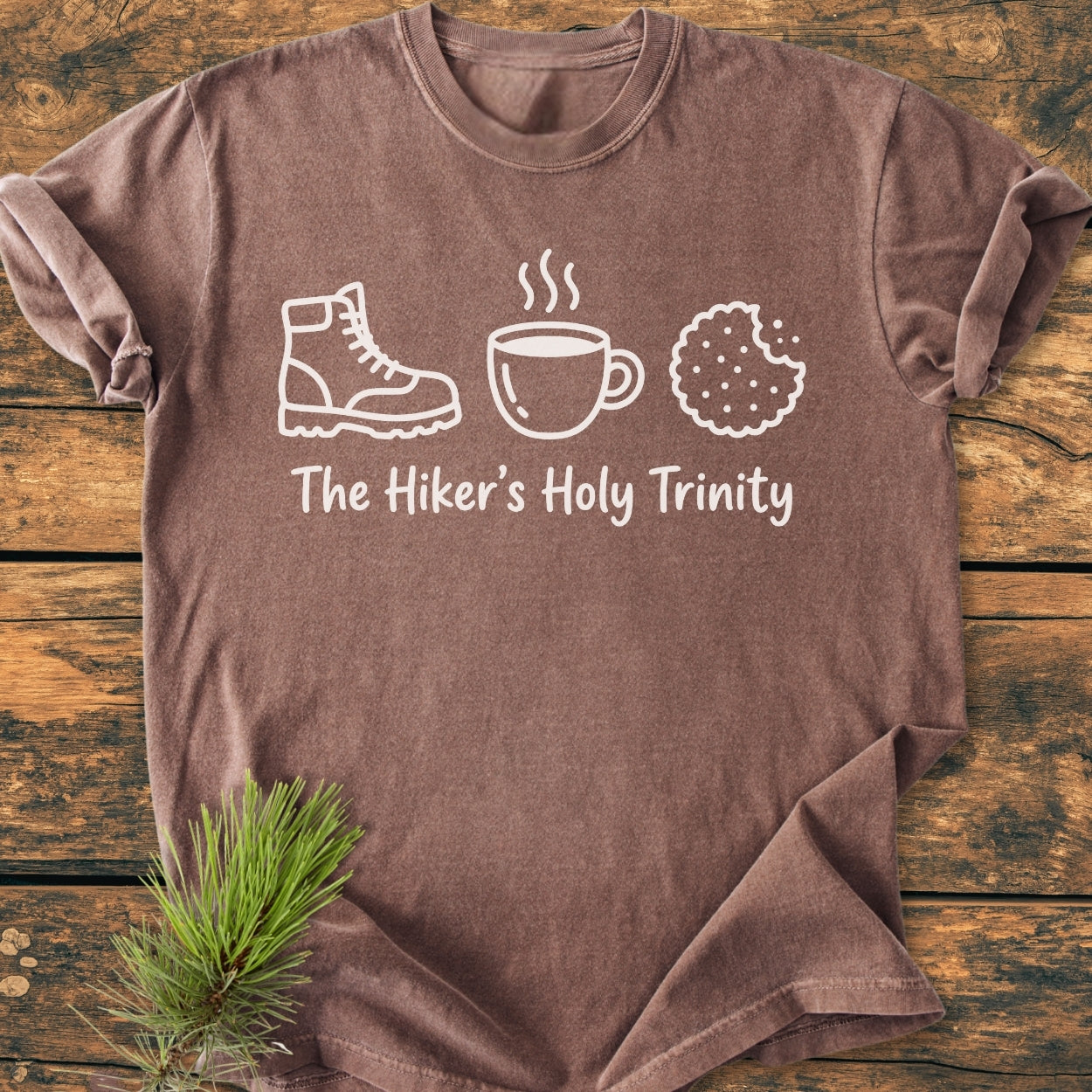 The Hiker's Holy Trinity - Vintage Wash Tee features white line-drawings of a boot, mug, and cookie on the chest, laid on wood with a pine sprig, highlighting comfort and durability.