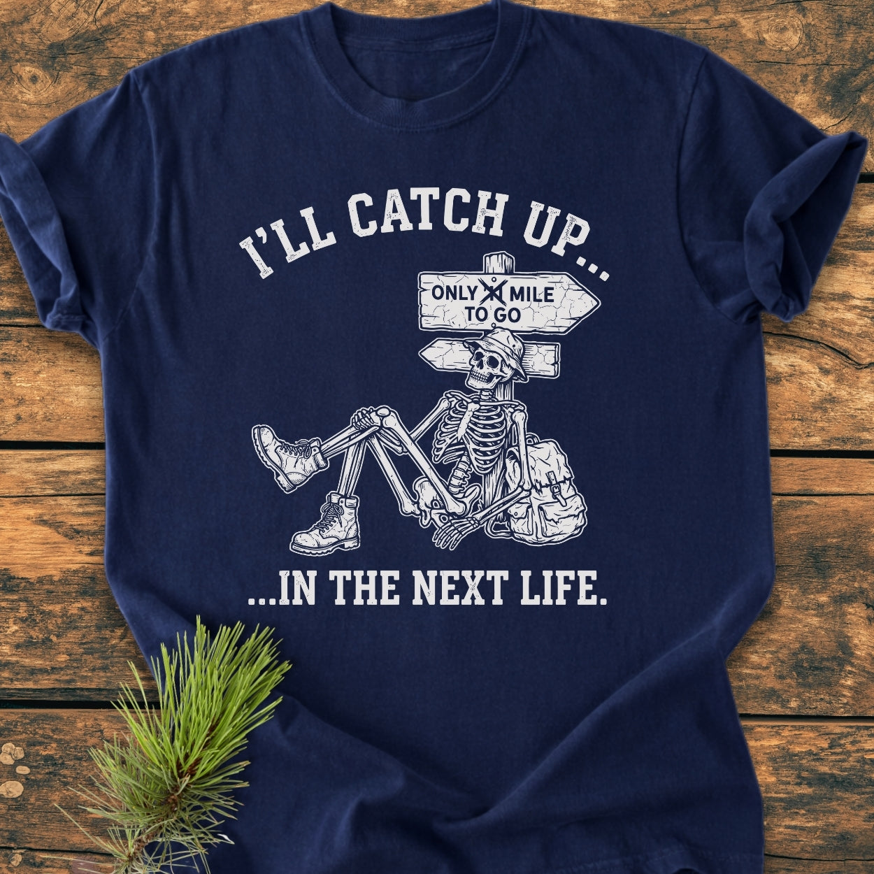 I'll Catch Up - Vintage Wash Tee featuring a sketch of a skeleton in hiking attire resting against a signpost, printed on a navy-blue, garment-dyed shirt laid on wood.