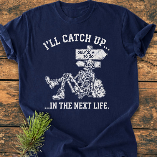 I'll Catch Up - Vintage Wash Tee featuring a sketch of a skeleton in hiking attire resting against a signpost, printed on a navy-blue, garment-dyed shirt laid on wood.