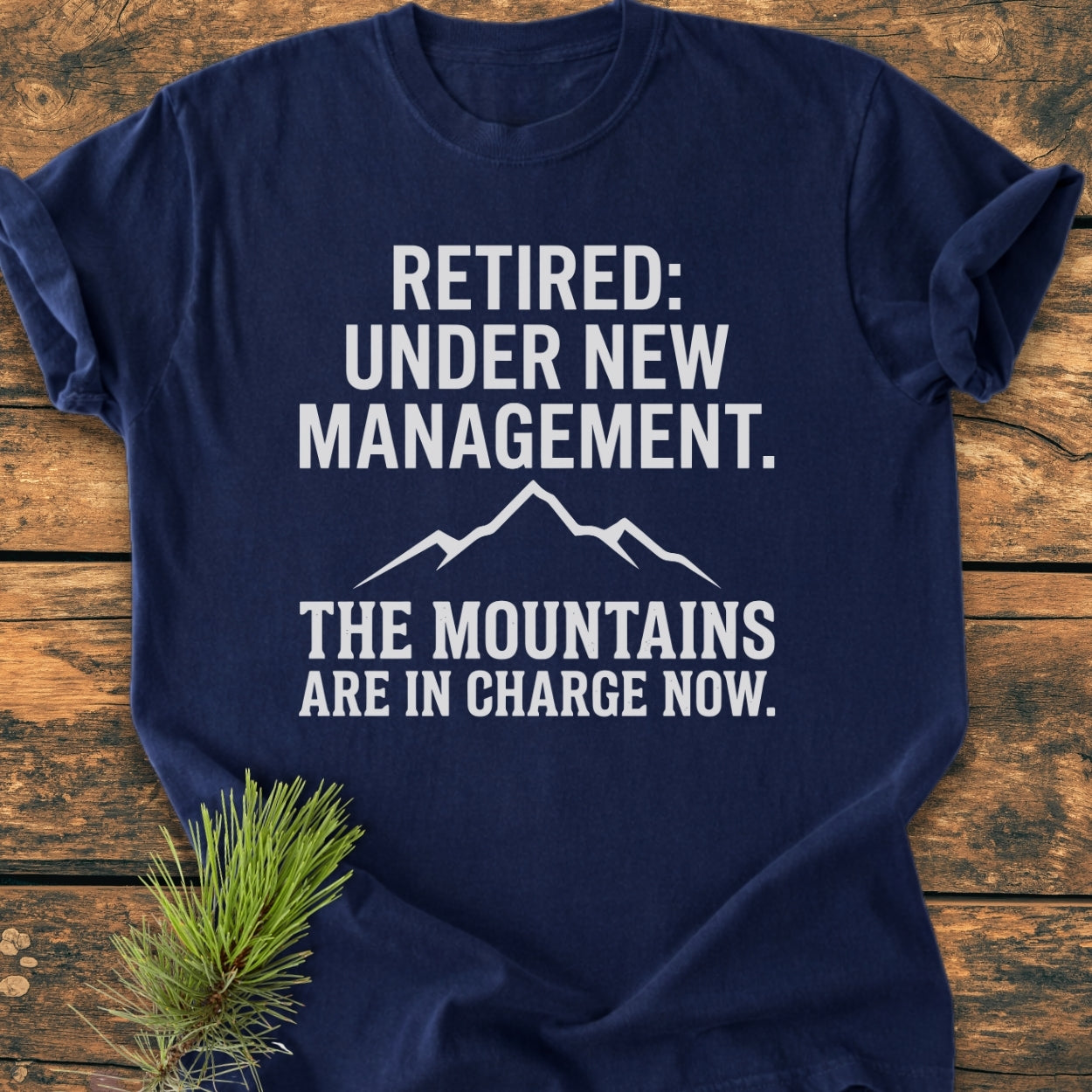 New Management - Vintage Wash Tee on wooden surface with pine sprig; navy-blue with white mountain graphic and text Retired: Under New Management. The Mountains Are In Charge Now. Relaxed, durable fit.