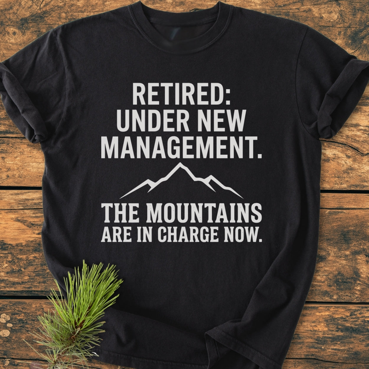 New Management - Vintage Wash Tee featuring white text RETIRED: UNDER NEW MANAGEMENT and THE MOUNTAINS ARE IN CHARGE NOW with a mountain graphic, laid on wood with a pine sprig detail.