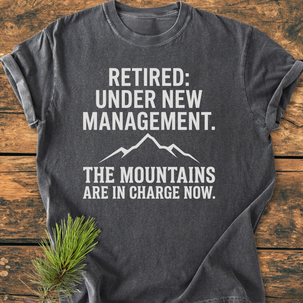 New Management - Vintage Wash Tee: Charcoal-gray shirt with white lettering RETIRED: UNDER NEW MANAGEMENT and mountain graphic, displayed on wood with a pine sprig. Comfortable, durable 100% ring-spun cotton.