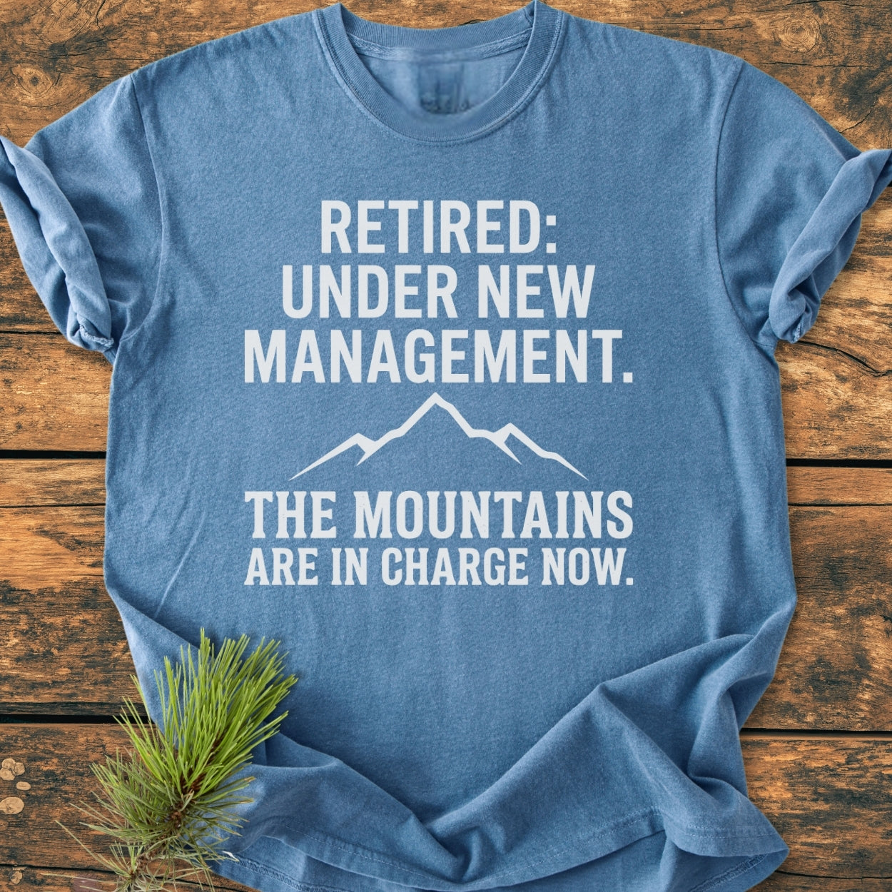 New Management - Vintage Wash Tee featuring a mountain graphic and humorous retirement text, displayed on a wooden surface with rolled sleeves and a pine sprig, emphasizing its rustic, outdoorsy appeal.