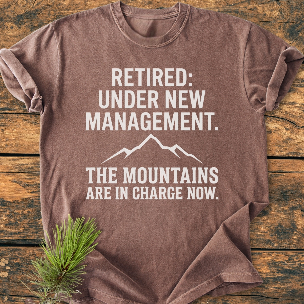 New Management - Vintage Wash Tee featuring a bold slogan and mountain graphic on a brown short-sleeve, laid flat with pine sprig, emphasizing an outdoorsy, retirement theme.