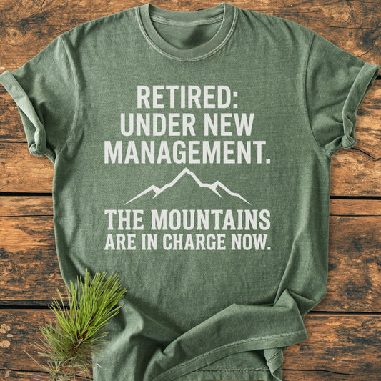 New Management - Vintage Wash Tee: Green heather T-shirt with bold white lettering and a mountain silhouette, laid on wood. A pine sprig complements its outdoorsy theme, emphasizing comfort and relaxed fit.