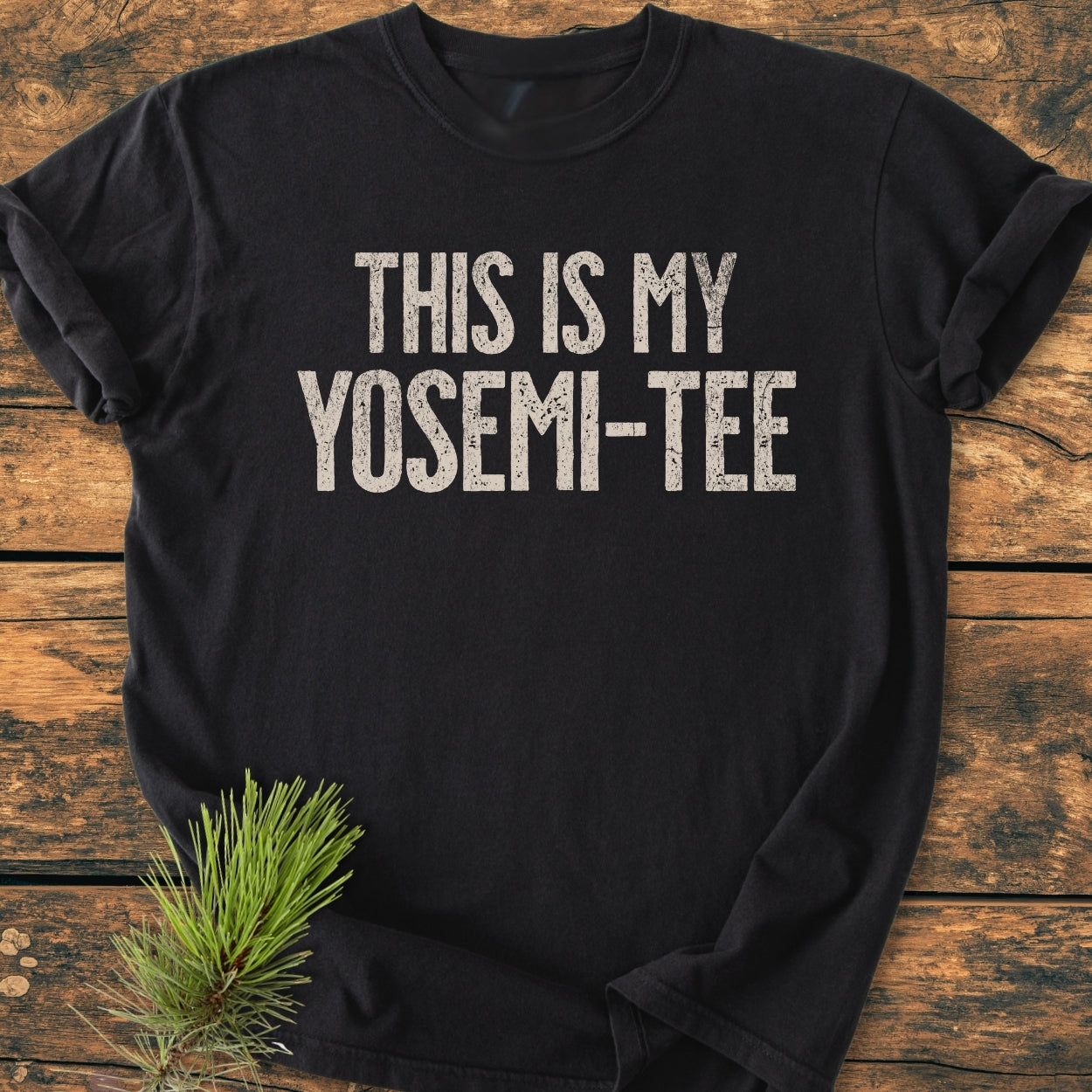 “This Is My Yosemi-Tee - Vintage Wash Tee” displayed on rustic wood, features distressed white lettering on a black background. Pine needles add an outdoorsy touch, emphasizing its relaxed, durable design.