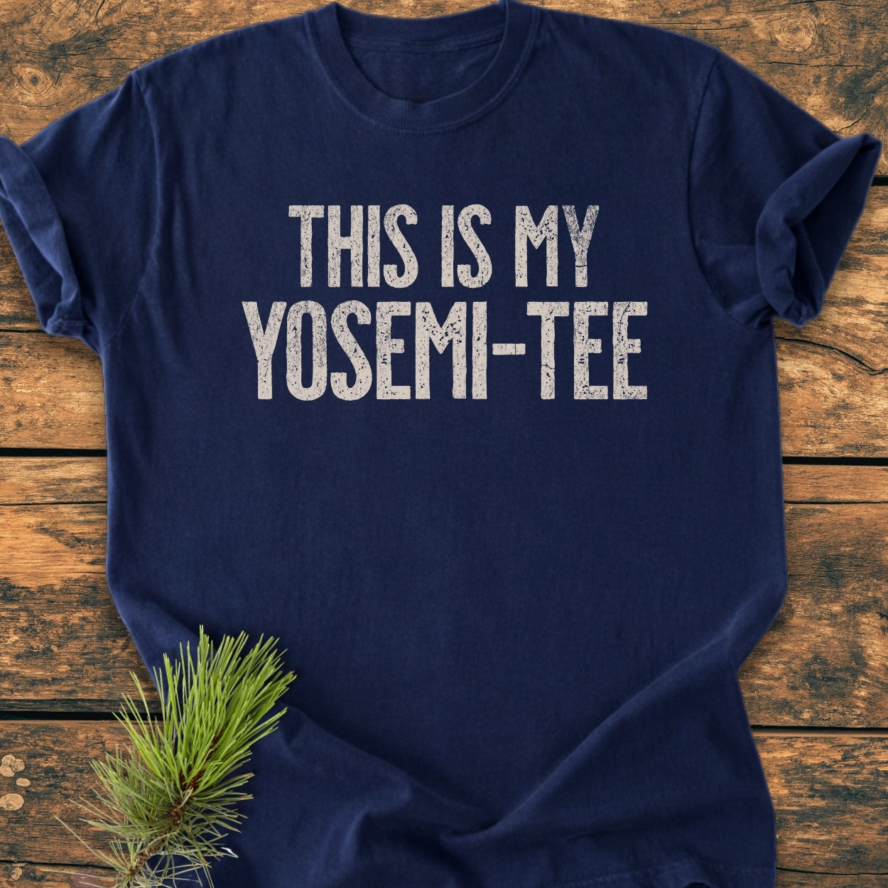 This Is My Yosemi-Tee - Vintage Wash Tee features a navy-blue color with a distressed white print on the chest, laid on wood. A pine bough accentuates the rustic display.