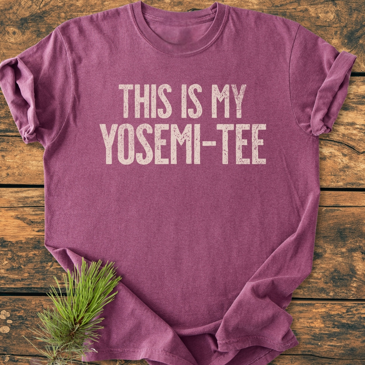 Flat-lay of This Is My Yosemi-Tee - Vintage Wash Tee, showcasing a distressed print on a heathered maroon T-shirt, styled with a pine needle sprig on a wooden surface.