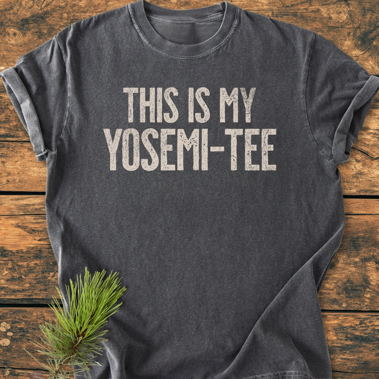 Charcoal-gray This Is My Yosemi-Tee - Vintage Wash Tee laid flat, featuring distressed white lettering across the chest on a rustic surface with a pine sprig accenting the scene.
