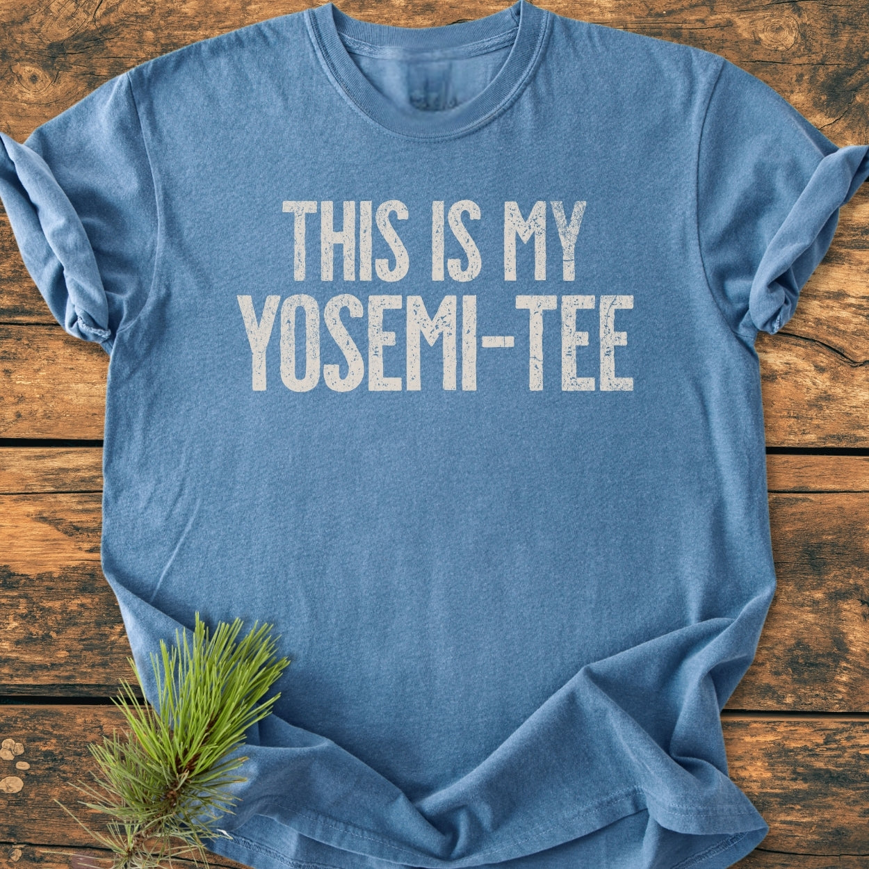 This Is My Yosemi-Tee - Vintage Wash Tee shown flat with rolled sleeves on a wooden surface, featuring distressed white lettering. A pine sprig adds an outdoorsy touch, highlighting its national-park theme.