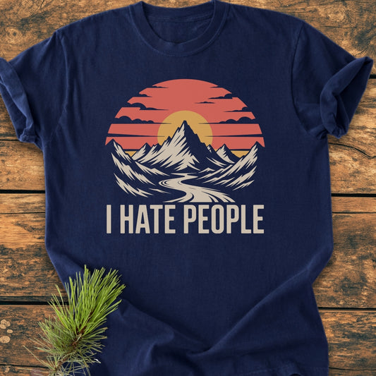 I Hate People - Vintage Wash Tee features a mountain scene with peaks and a sun/moon on a navy T-shirt, laid on wood, with a pine branch accenting the display.