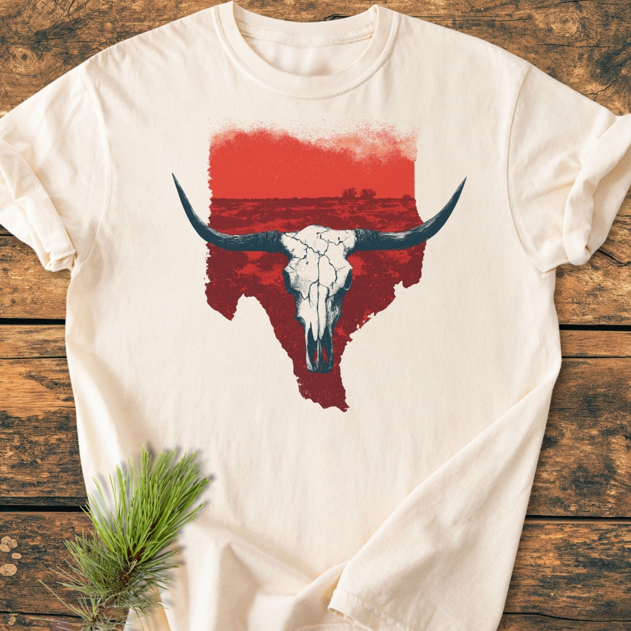 Hike Texas - Vintage Wash Tee featuring a longhorn skull graphic on a cream T-shirt, displayed on a wooden surface with a pine sprig accent, highlighting its relaxed, durable design.