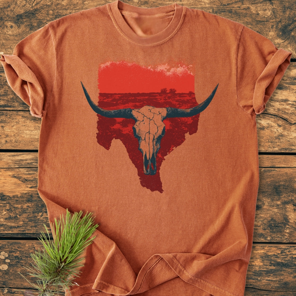 Hike Texas - Vintage Wash Tee features a rust-orange color, rolled sleeves, and a large steer skull graphic on the front, laid on a wooden surface with a small pine sprig beside it.