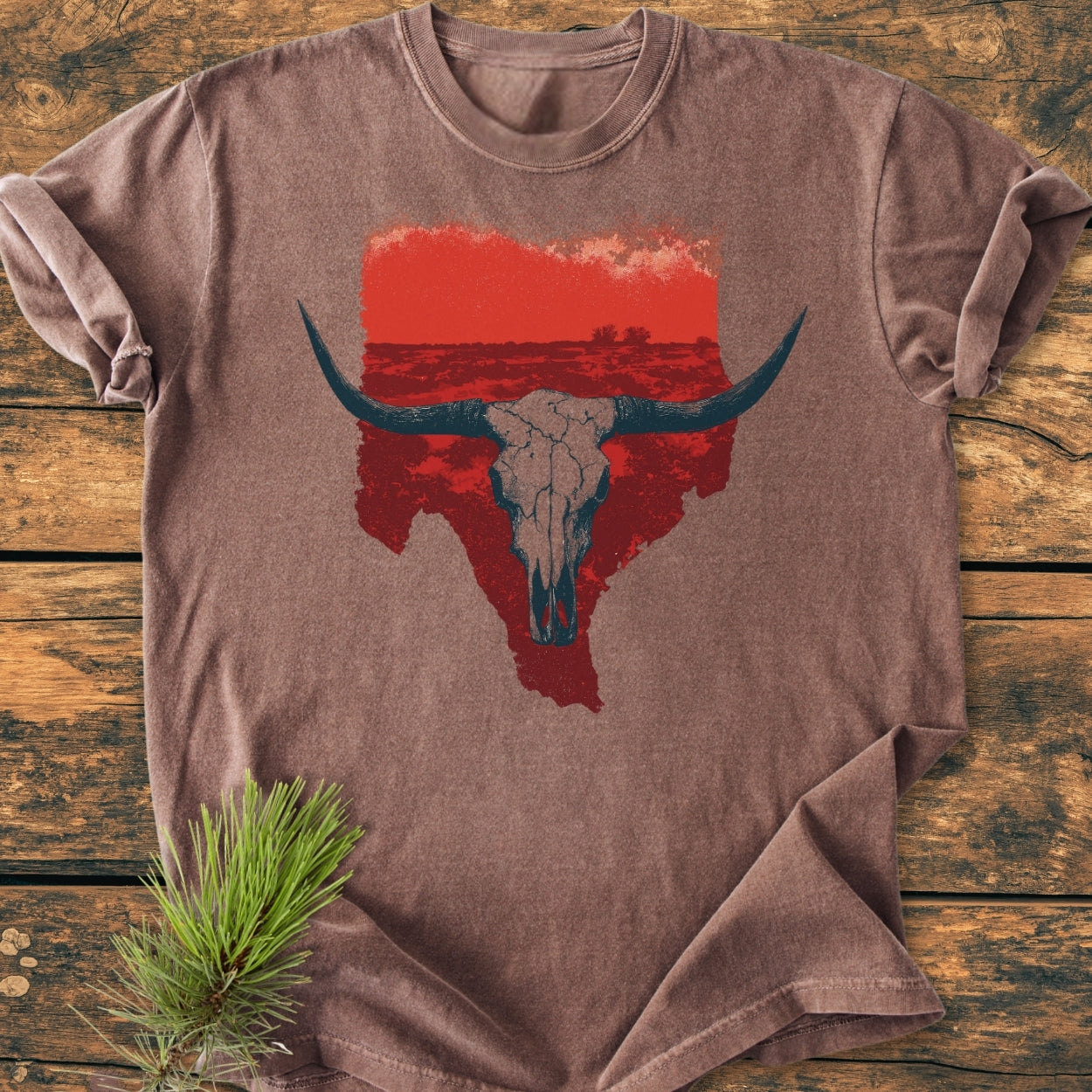 Hike Texas - Vintage Wash Tee: Brownish t-shirt with a red Texas silhouette and longhorn skull graphic, laid on wood. Rustic style enhanced by a pine sprig, showcasing the tee's relaxed fit and durability.