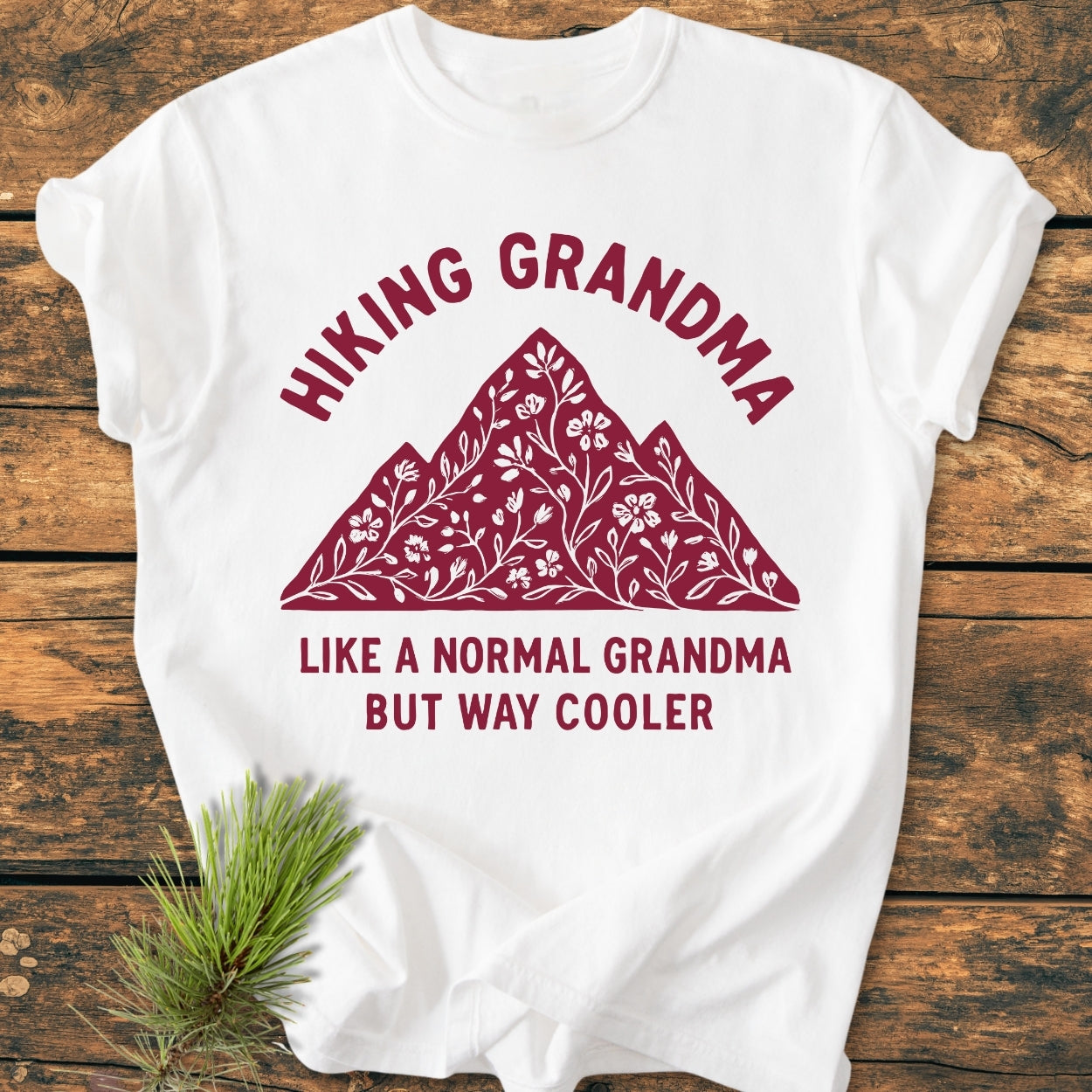 White Hiking Grandma Floral - Vintage Wash Tee with a stylized mountain graphic and floral vine pattern, laid on wood with pine sprig. Features slogan about being a cooler grandma.