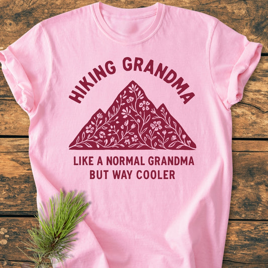 Hiking Grandma Floral - Vintage Wash Tee features a mountain silhouette with floral motifs and playful text, laid flat on a wooden surface, showcasing its relaxed fit and durable stitching.
