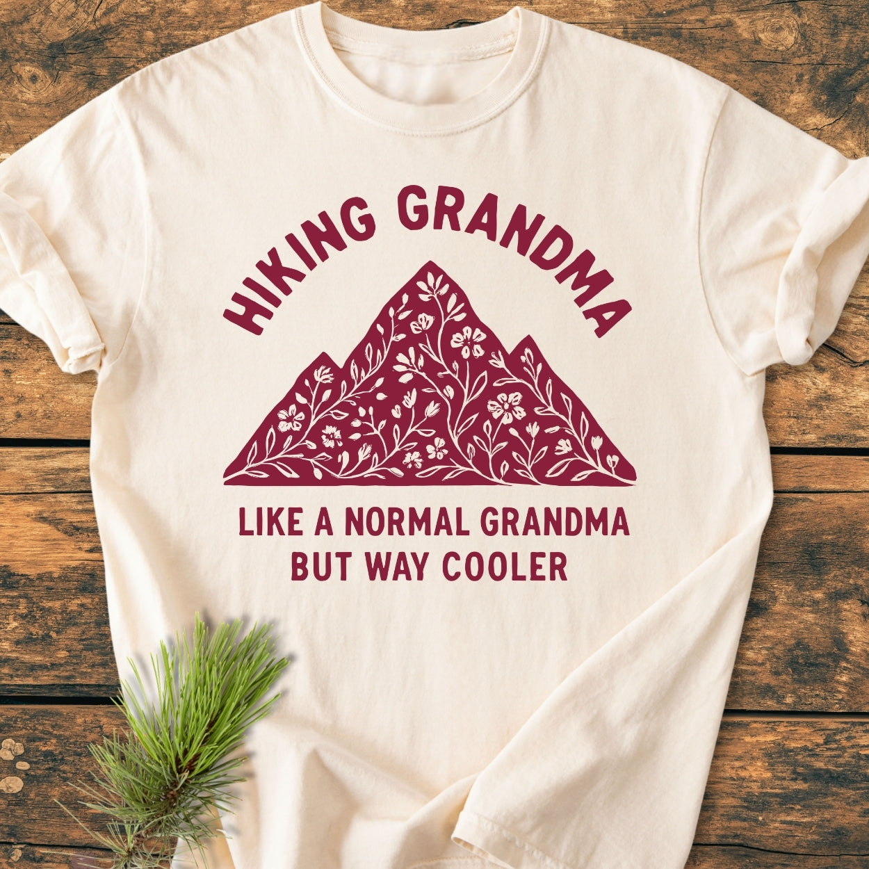 Hiking Grandma Floral - Vintage Wash Tee features a maroon mountain design with floral motifs and playful text. Displayed on wood, a pine sprig enhances its cozy, durable appeal.