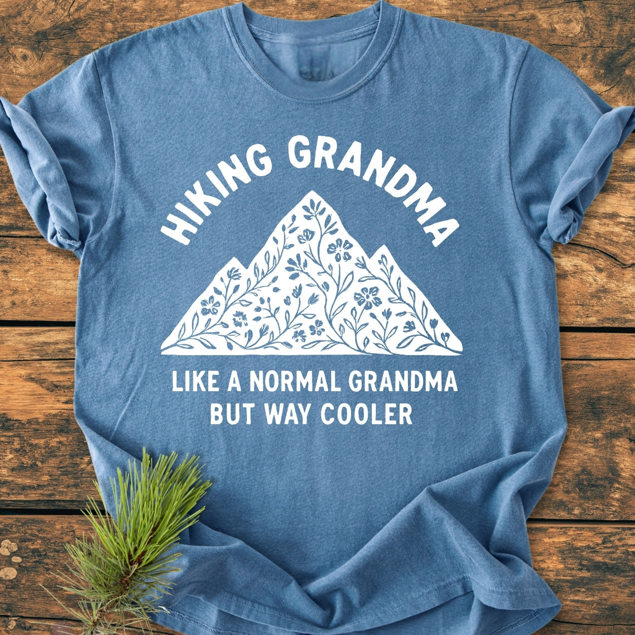 Hiking Grandma Floral - Vintage Wash Tee features a mountain with floral motifs and playful text. Laid on wood with pine sprig, it's a cozy, relaxed-fit shirt made of soft, durable cotton.