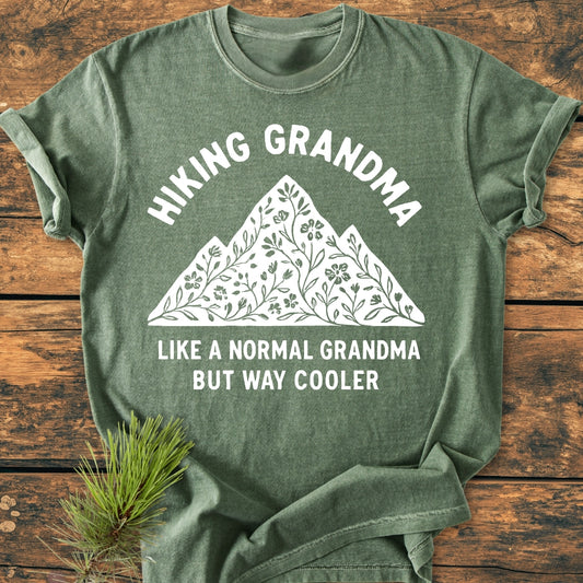 Hiking Grandma Floral - Vintage Wash Tee features a heather-green shade with mountain and floral motifs, bold white text, and relaxed fit, displayed on a wood surface with a pine sprig accent.