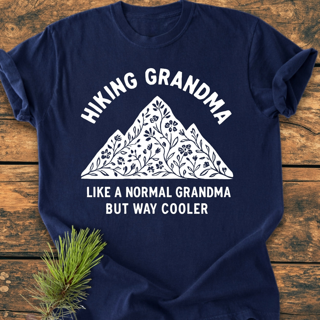 Navy-blue Hiking Grandma Floral - Vintage Wash Tee with a mountain and floral graphic, text above and below, laid on wood with a pine sprig accentuating its rustic appeal.