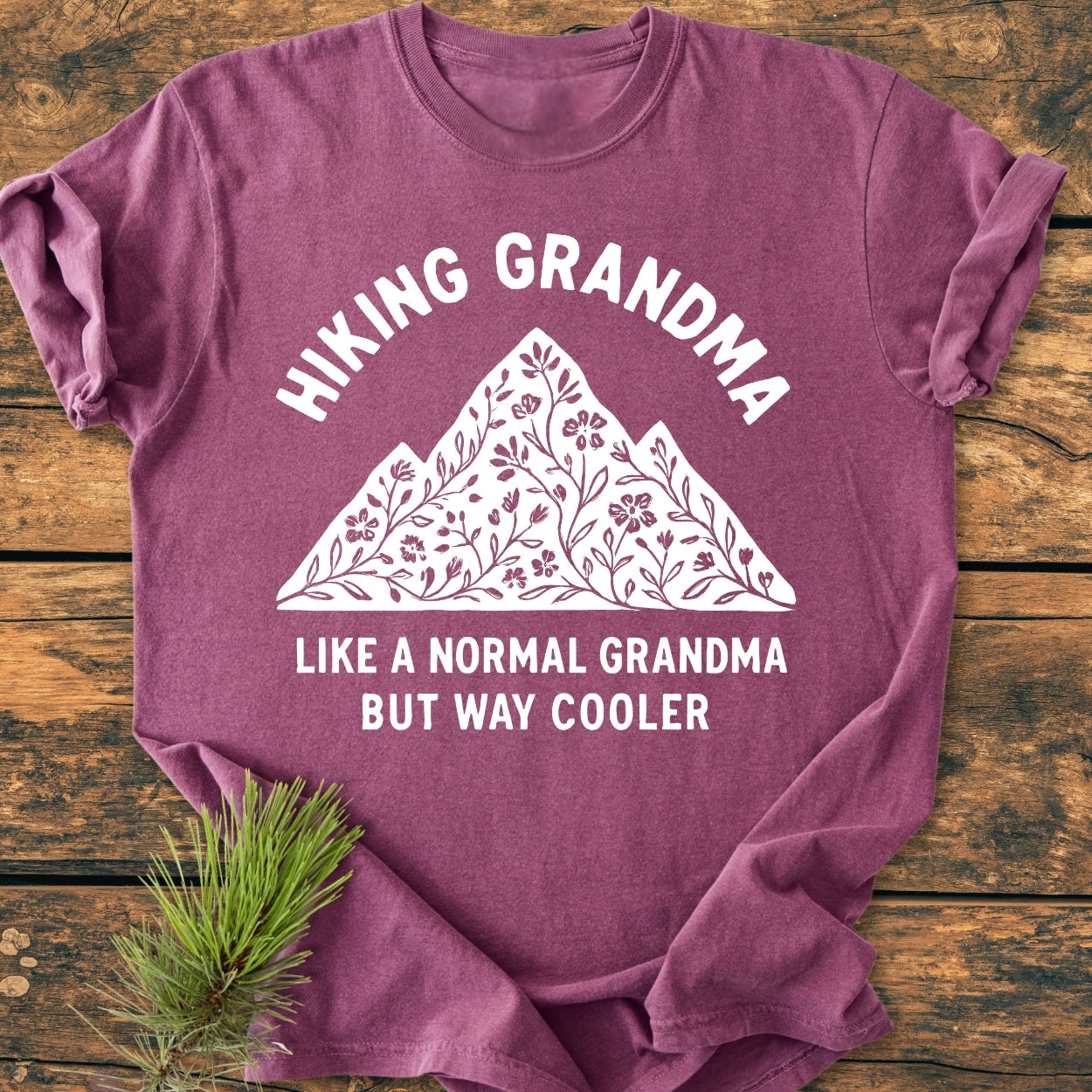 Hiking Grandma Floral - Vintage Wash Tee featuring a stylized mountain and floral graphic, laid flat with pine sprig. Made from soft, durable, garment-dyed 100% cotton, offering a relaxed fit.