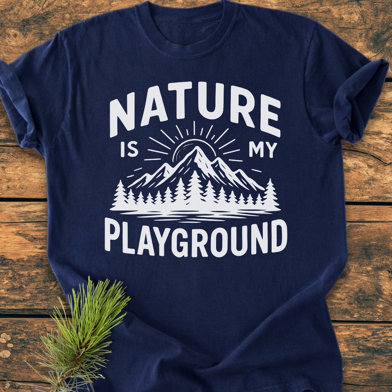 Nature Is My Playground - Vintage Wash Tee: Navy-blue tee featuring a mountain graphic with text, laid flat with a pine branch prop, showcasing its relaxed fit and soft, garment-dyed fabric.