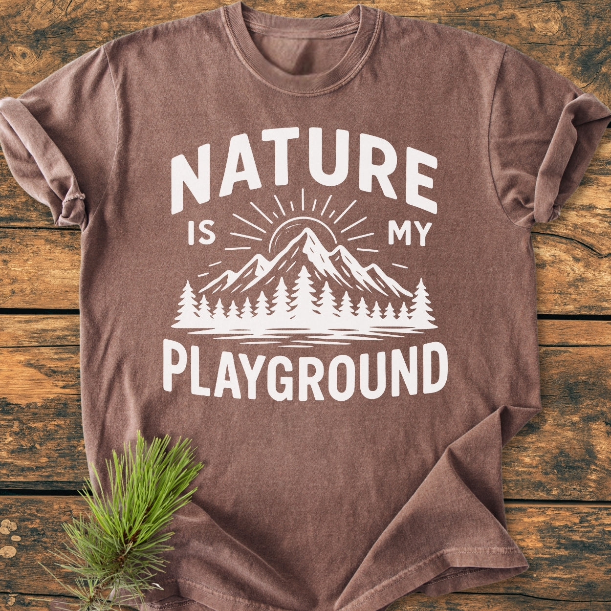 Nature Is My Playground - Vintage Wash Tee featuring a mountain and sun graphic with pine trees, laid flat on wood. Crafted from soft, garment-dyed cotton for comfort and durability.