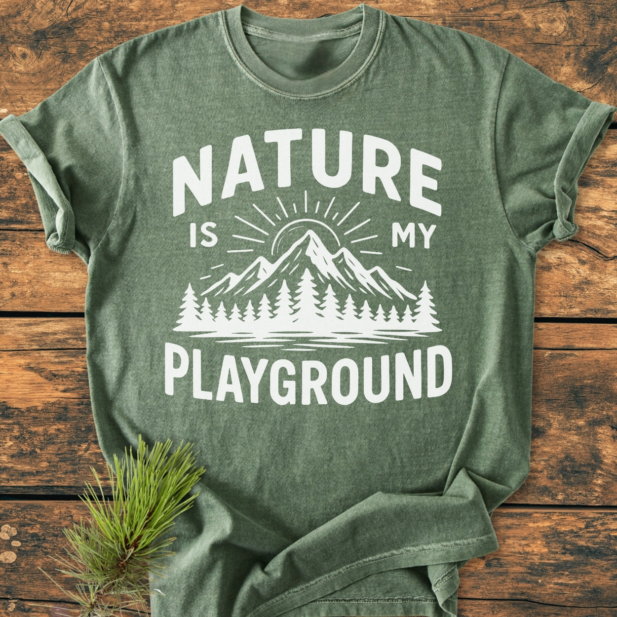 Nature Is My Playground - Vintage Wash Tee, featuring a white mountain and sunbeam graphic with the slogan, laid flat on wood, paired with a pine sprig for a natural touch.