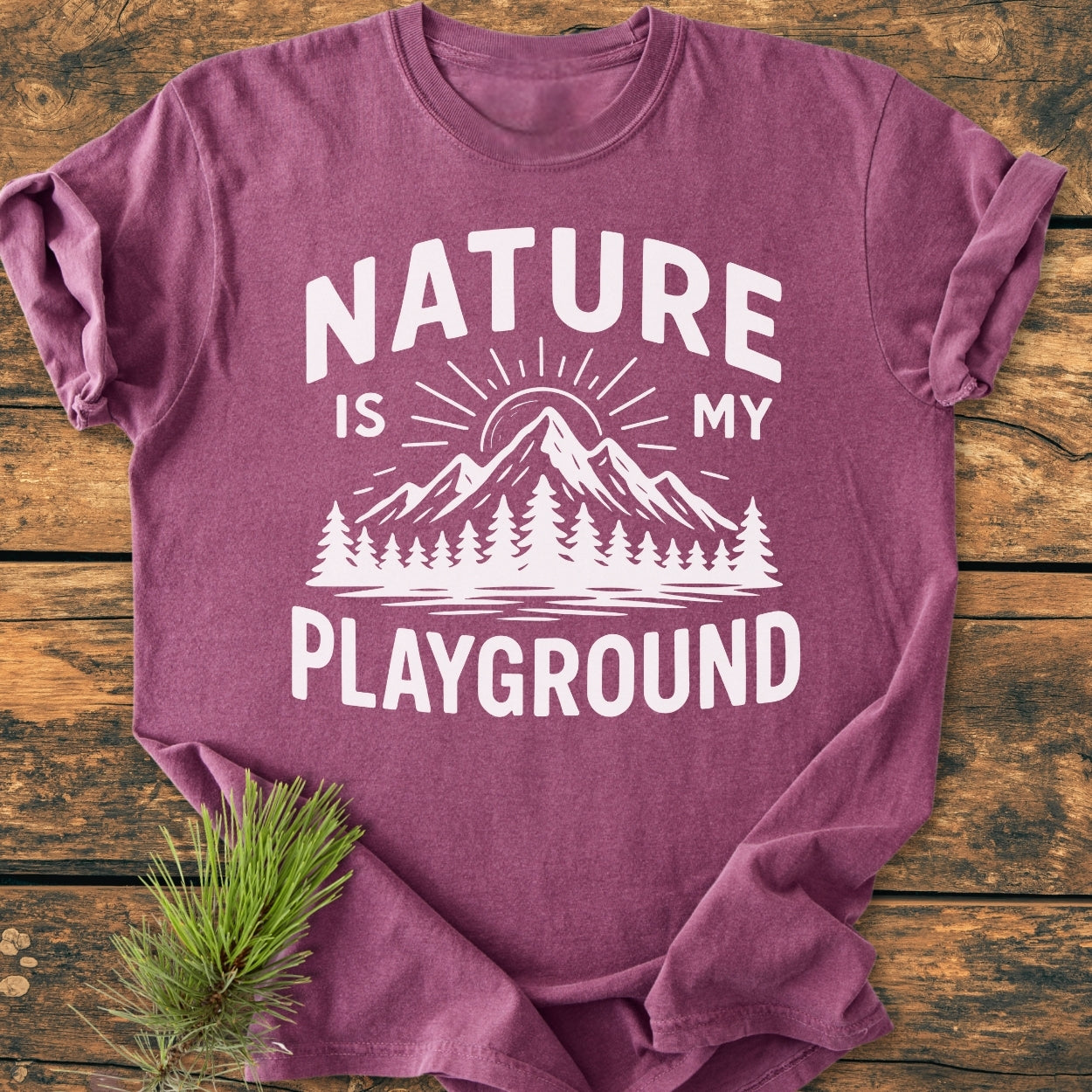 Nature Is My Playground - Vintage Wash Tee, a heathered purple shirt with a mountain and pine-tree print, laid on rustic wood, showcasing its soft-washed, durable fabric and relaxed fit, with a pine sprig detail.