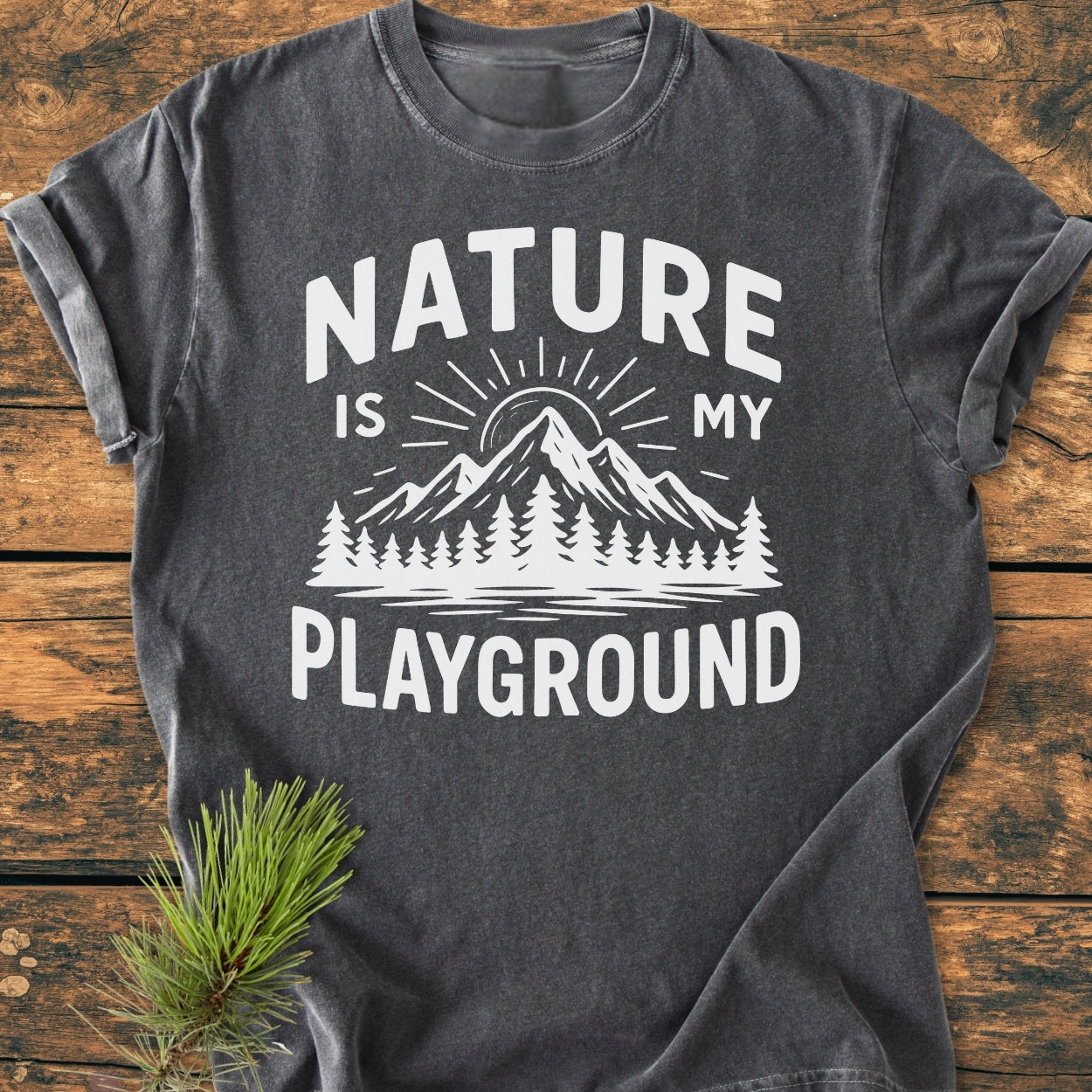 Nature Is My Playground - Vintage Wash Tee features a charcoal-gray design with white lettering and a nature-themed graphic. Laid on wood with a pine sprig, showcasing its casual, durable style.